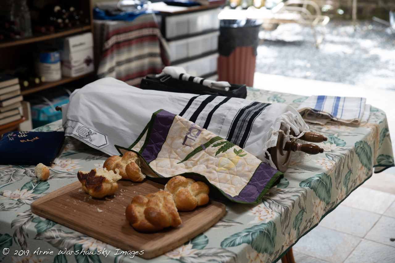 A table with two loaves of challah on a partially covered board, next to a covered Torah scroll.